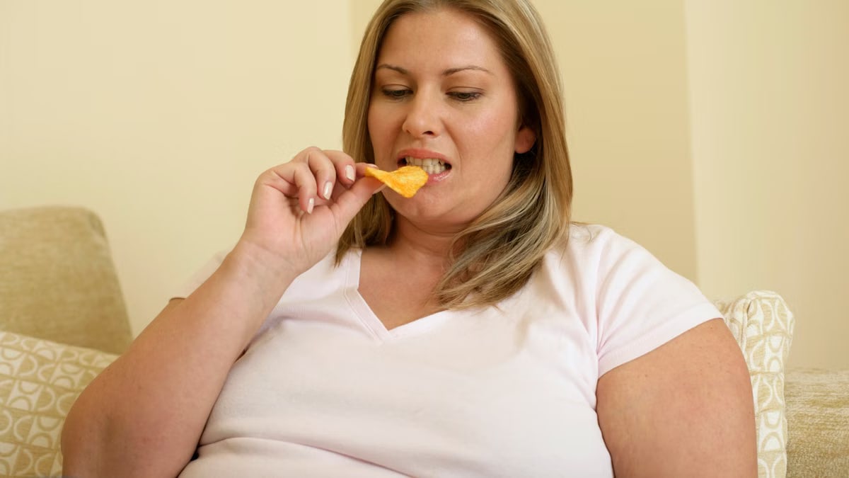 A woman sits comfortably on a couch indoors, enjoying a snack of potato chips.